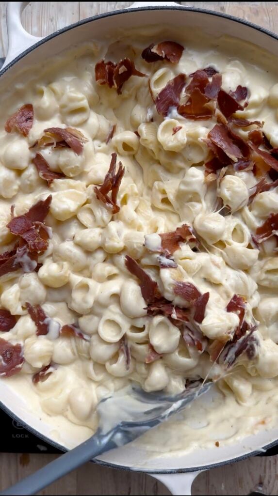 Noodles and crispy prosciutto being stirred into a creamy-cheese sauce.