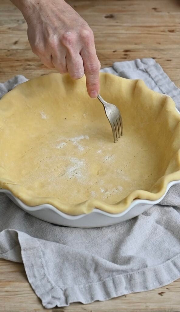 A hand docking a pie crust in a pie pan with a fork.