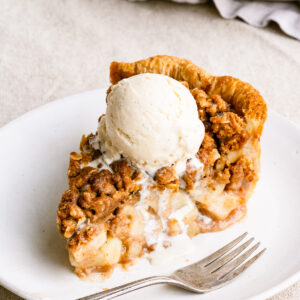 Slice of apple crumble pie topped with melting ice cream on a plate next to a fork.