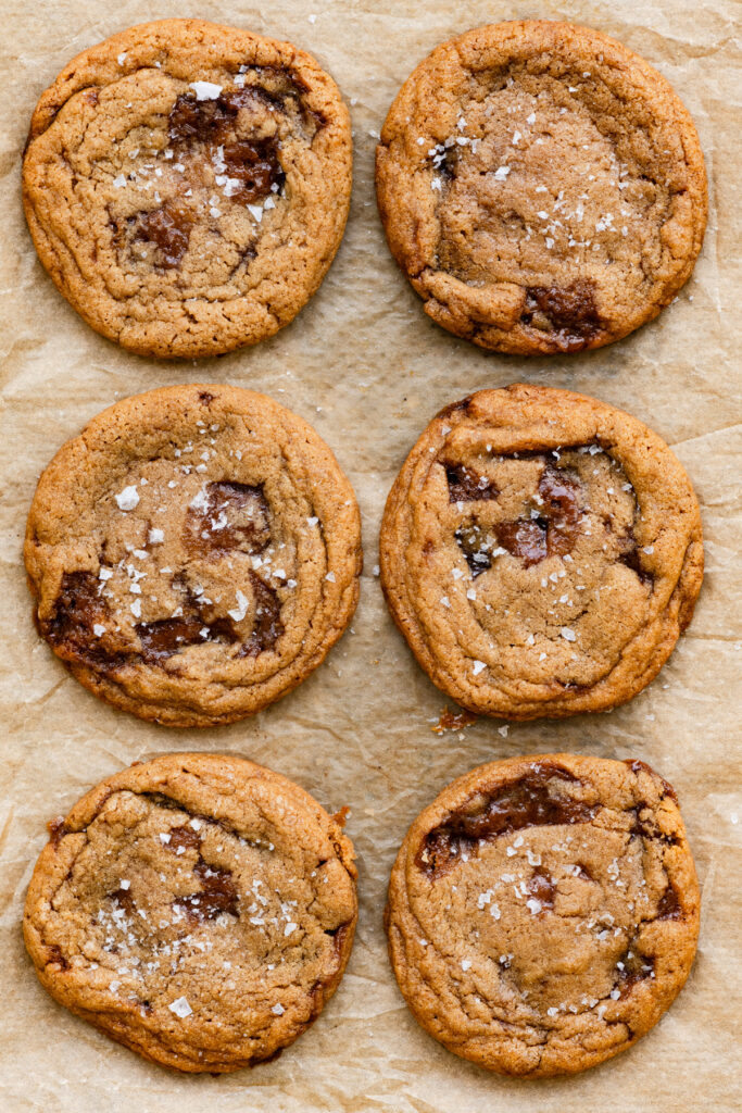 Six large cookies baked with chunks of melted toffee in them on a parchment paper lined tray.