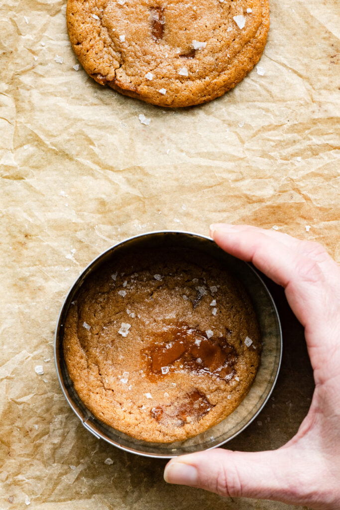 A hand reshaping a cookie with a metal ring on a baking tray.