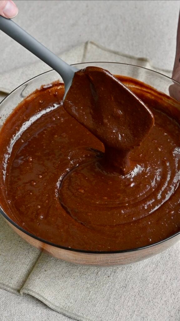 A spatula spooning out dark chocolate cake batter from a mixing bowl.