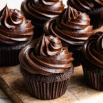 Five chocolate cupcakes topped with chocolate frosting on a wooden cuttingboard.