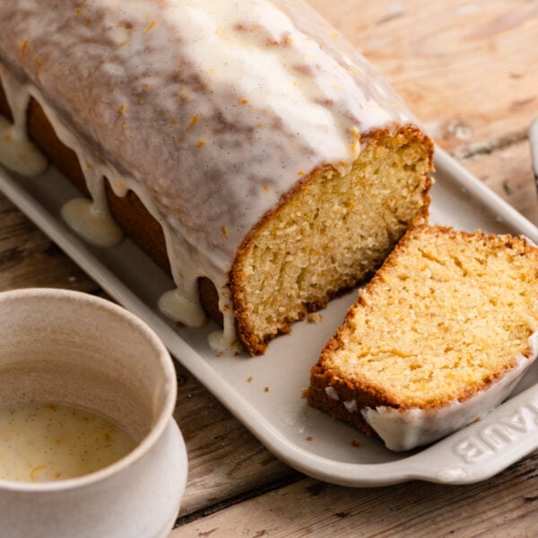 A cup of cake glaze next to a sliced cake topped with orange cake glaze.