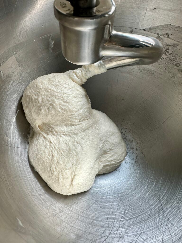 Pizza dough attached to a dough hook in the bowl of a stand mixer.