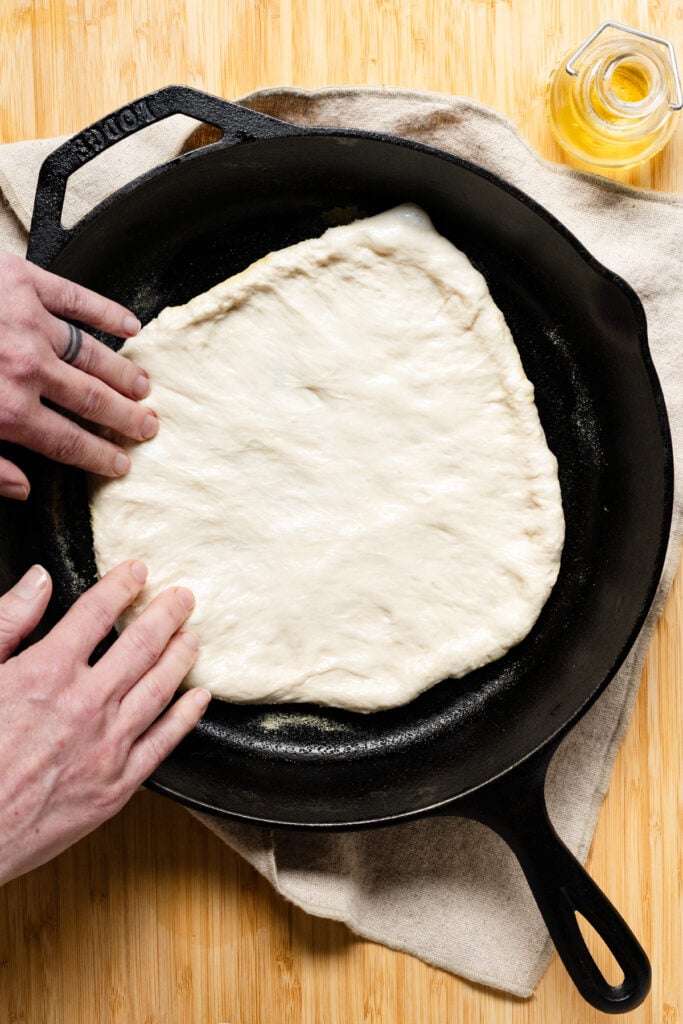 Two hands stretching pizza dough to cover the bottom of a cast iron skillet.
