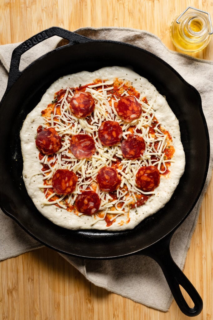 Overhead of pepperoni and cheese topped pizza dough in a cast iron skillet.