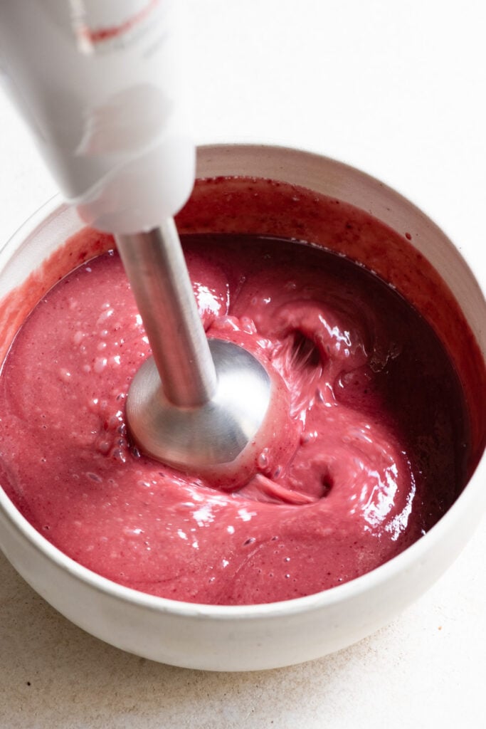 Immersion blender mixing liquid raspberry ganache in a bowl.