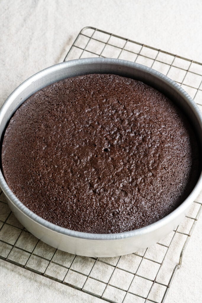 Baked chocolate cake inside a round cake pan on wire rack.