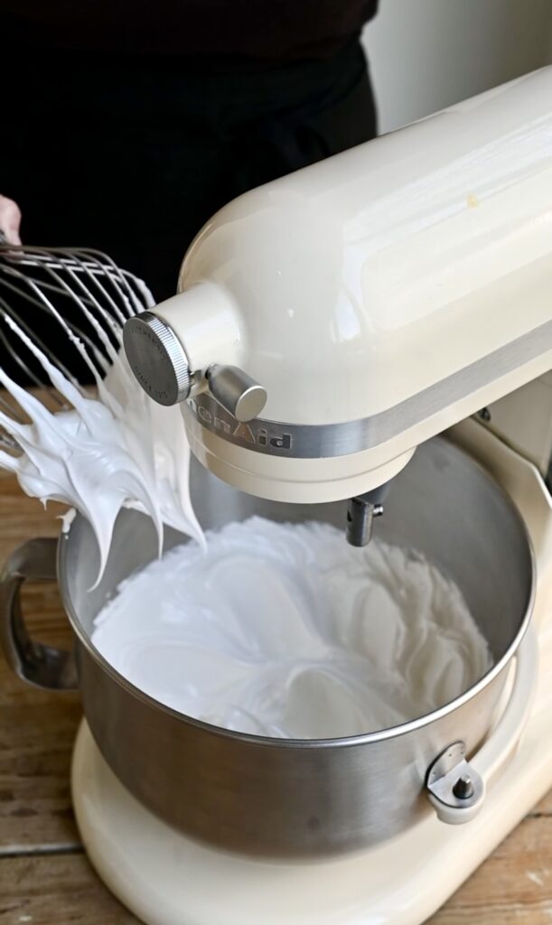 Whisk with stiff peak meringue pulling out of a mixing bowl.