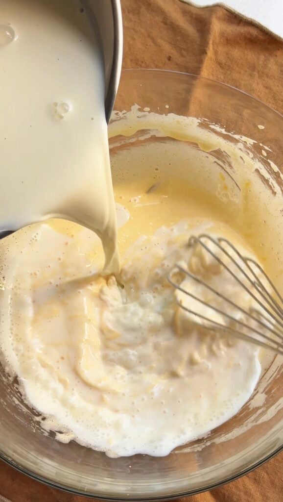 Pouring hot milk into a bowl of egg yolks with a whisk.
