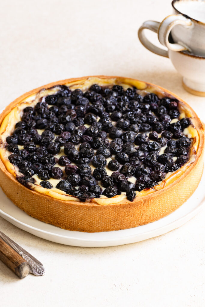 A blueberry French flan tart on a serving late next to coffee mugs and utensils.