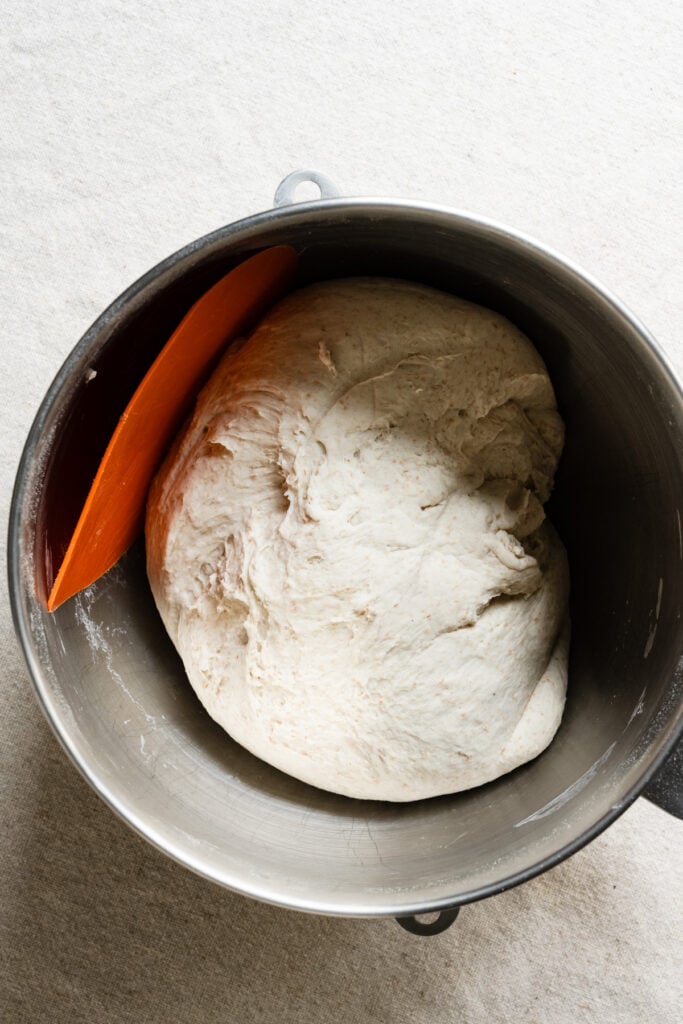 Overhead of bread dough in a mixing bowl with a bowl scraper.