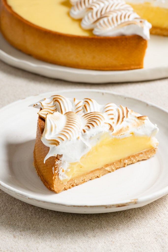Slice of lemon curd tart on a plate next to the full sized tart.