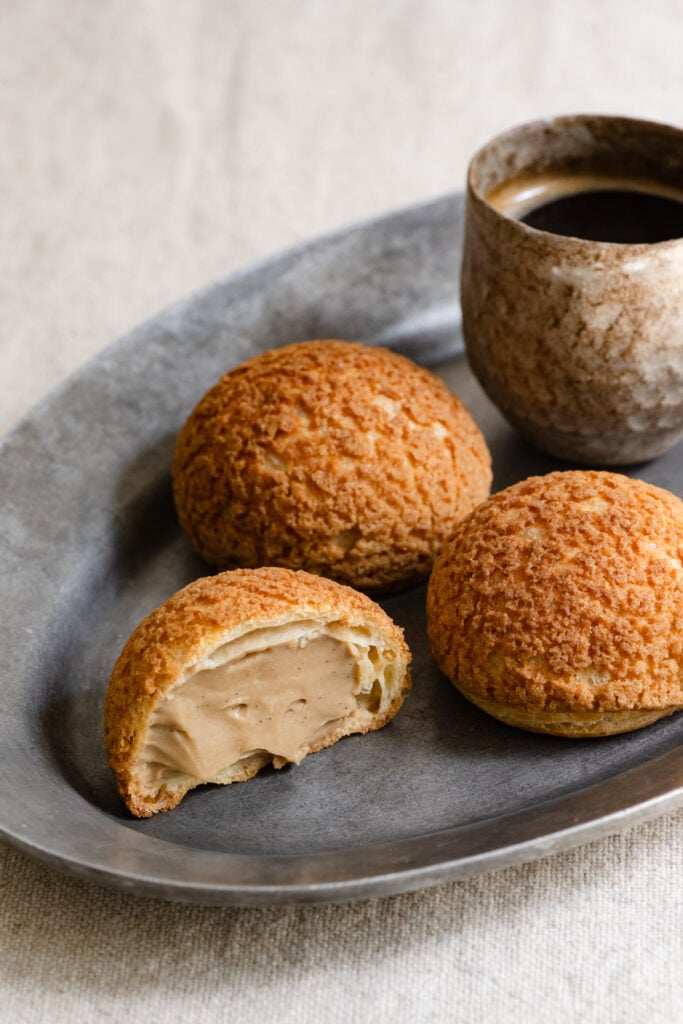 Two coffee cream puffs and one sliced in half on a plate with a cup of coffee.