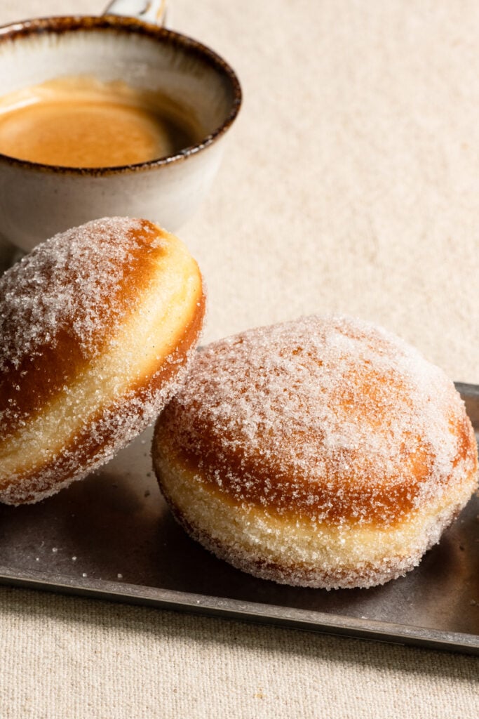 Two sugar coated brioche donuts on a plate next to a cup of coffee.