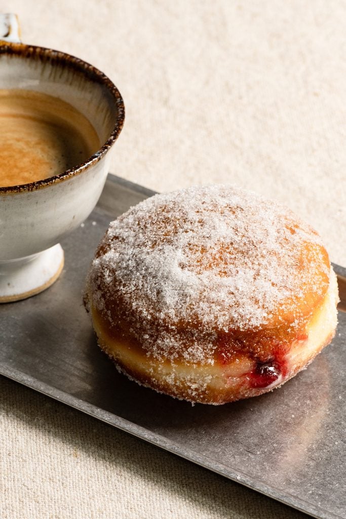 Raspberry jelly filled brioche donut on a plate next to a cup of coffee.